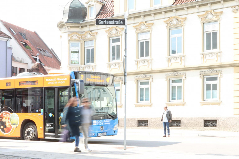 Ein Bus steht an der Haltestelle. Es sind unscharf Mesnchen zu sehen, die gerade ausgestiegen sind.